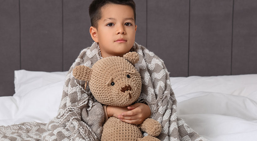 Symbolbild: Kranker Junge mit Teddybär sitzt auf dem Bett