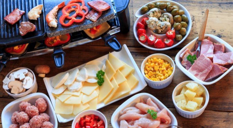 Raclette mit Schälchen, die mit verschiedenen Lebensmitteln gefüllt sind: z. B. Fleisch, Käse, Mais